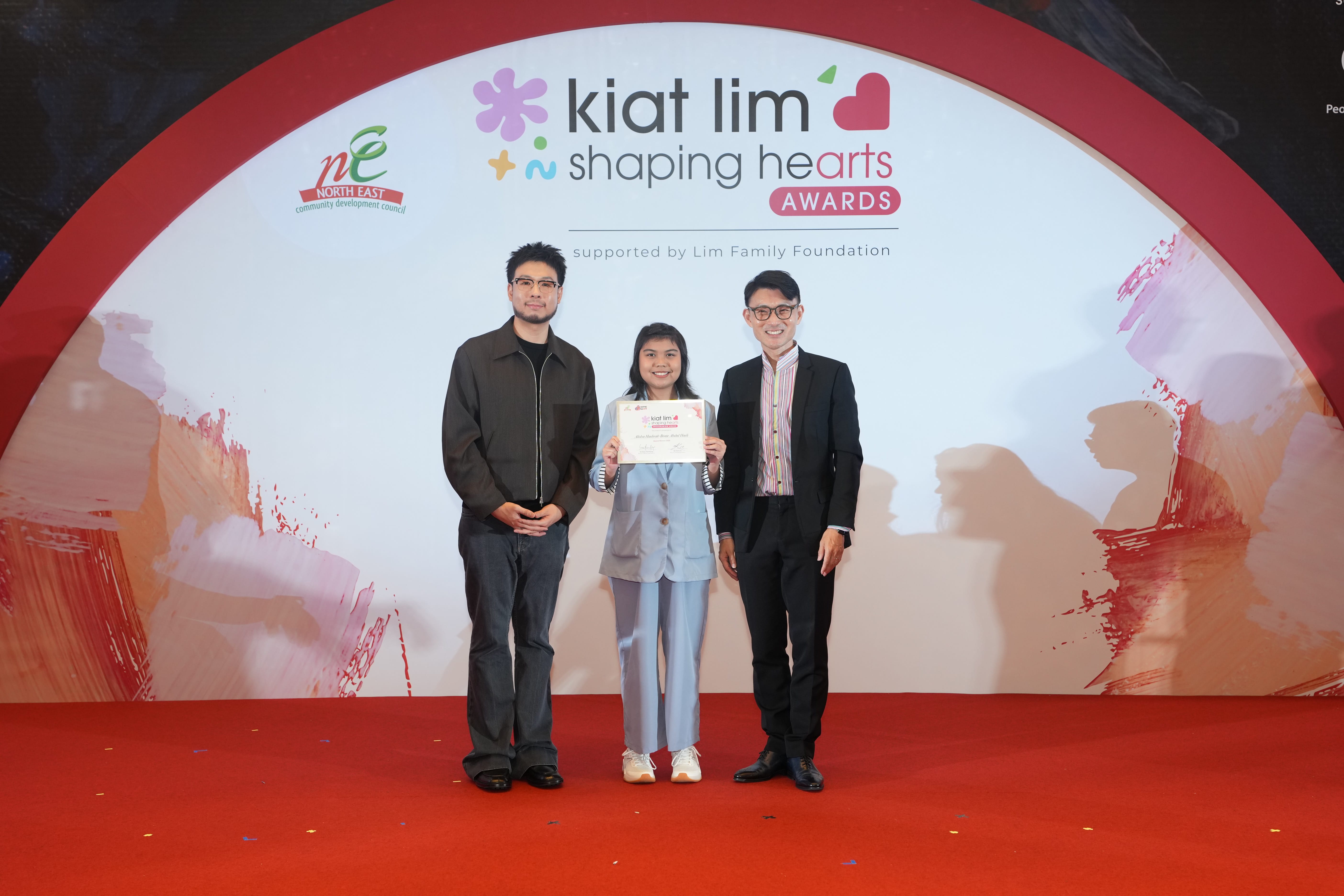 Award recipient standing on stage with the presenter, holding their certificate and posing for a group photo during the award ceremony, with a backdrop reading ‘Kiat Lim-Shaping Hearts Awards'.